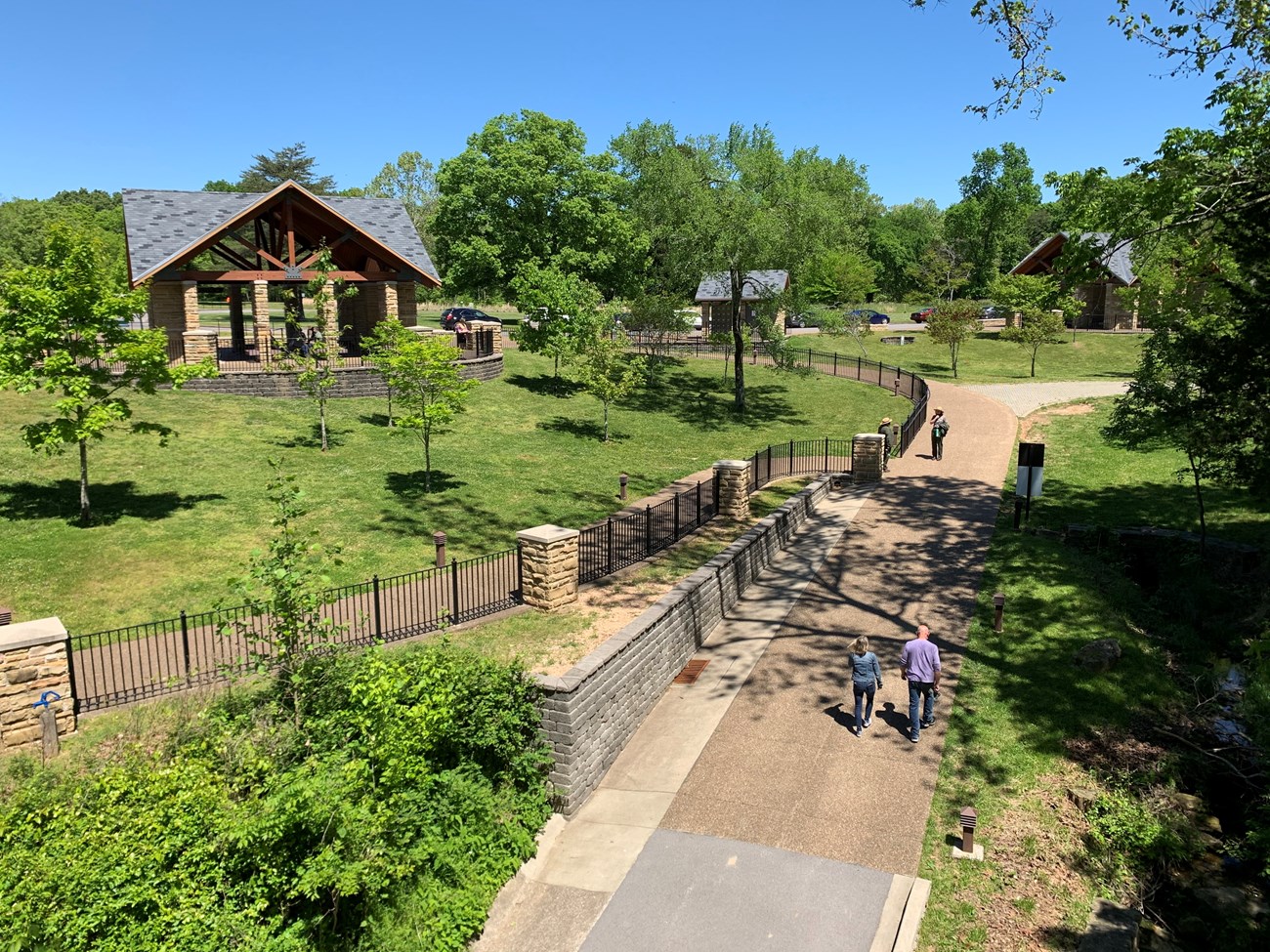 A high-angle view shows a visitor area including pathways, fencing, trees scattered in turf, and rustic, pavilion-like structures