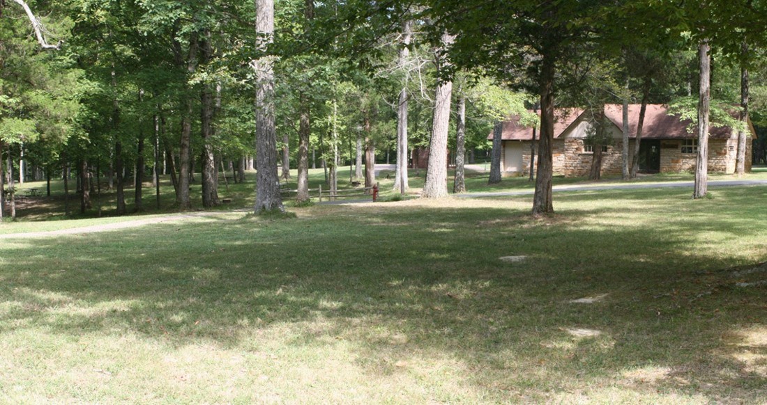 Tall, deciduous trees shade turf in front of a rustic-style, one-story building.