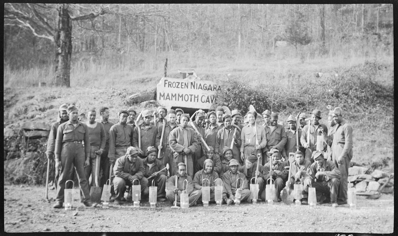 A group of young African American men with tools, lanterns, and work uniforms pose for a photo in front of the Frozen Niagara Mammoth Cave entrance
