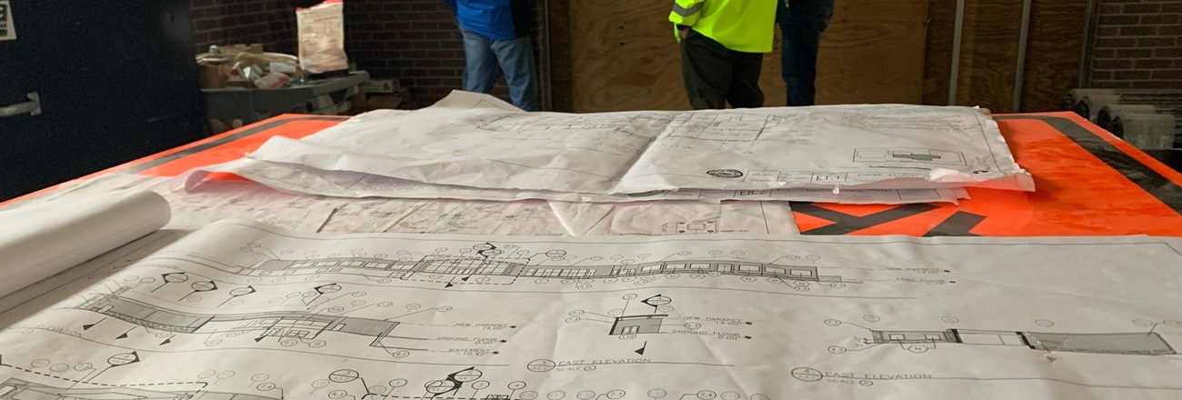 Large sheets of paper covered in architectural drawings, with three people talking in the background