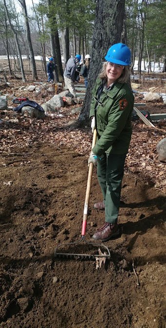 A woman in NPS uniform, hardhat, and gloves pulls a rake through dark soil