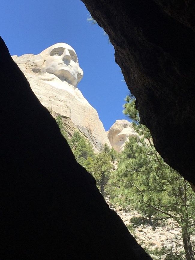 A view upwards towards the faces of the Mount Rushmore sculpture, seen through an opening between rocks along a trail.