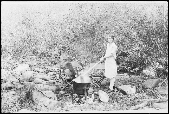 A woman uses a long stick to stir a steaming pot over an outdoor fire. A dog sits beside her on one of two camp chairs.