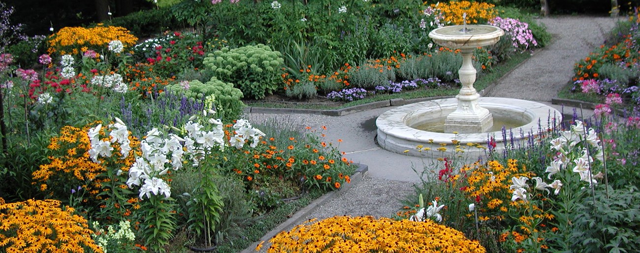 A round fountain on a pedestal stands where two paths cross in a garden of colorful flowershs cross in a garden