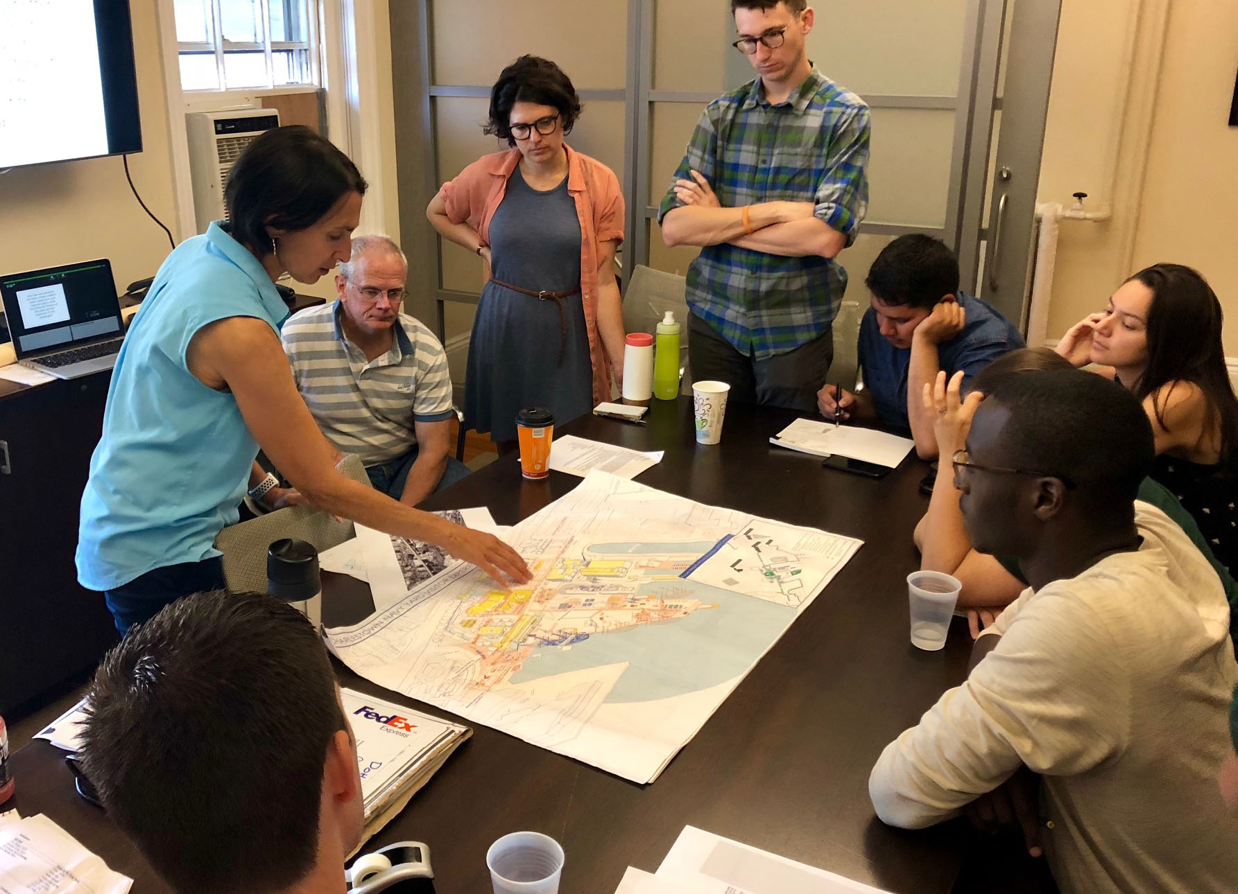 A group of people is gathered around a wooden table, looking on as one person points to a site plan.