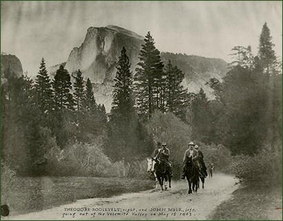 Historic image of four men on horses riding a trail, mountains and trees in the background.