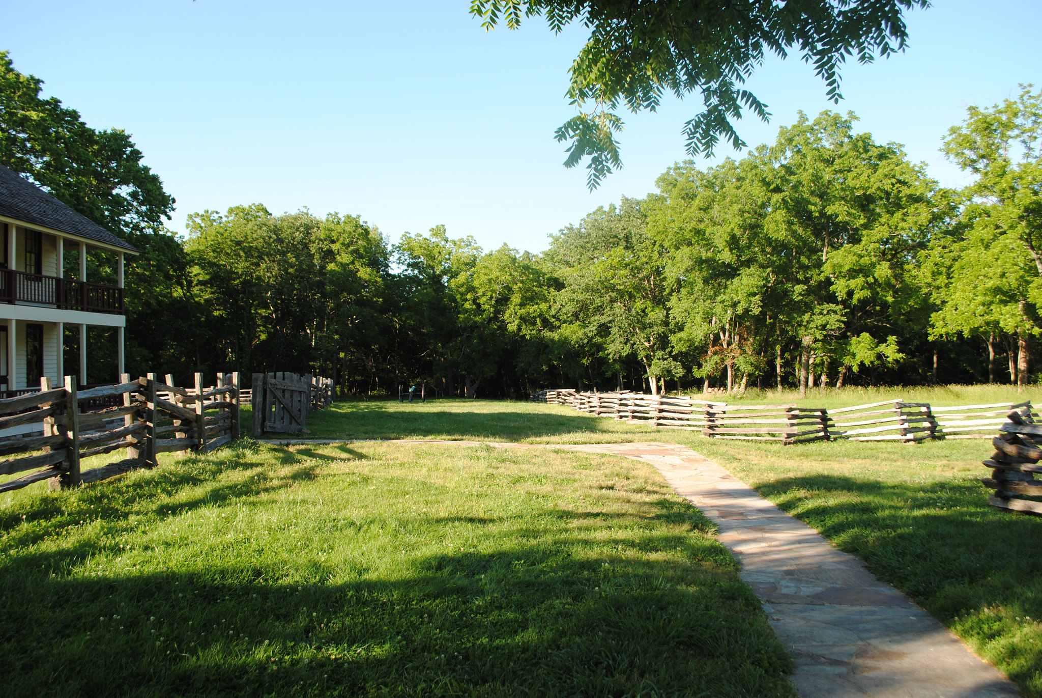 Wooden fences along a path through an open area of grass, with a two-story structure to the left and a border of leafy trees.