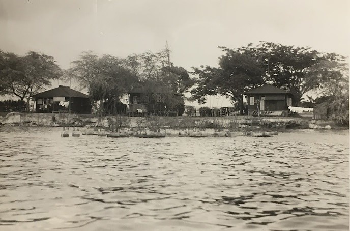 A view over water to a row of three one-story bungalows, surrounded by leafy trees.