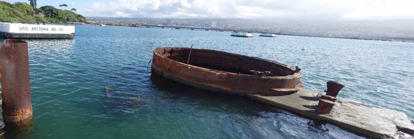 A view over greenish-blue water includes a round, metal section of the USS Arizona protruding from the water's surface. A series of mooring quays and distant land are visible in the background.