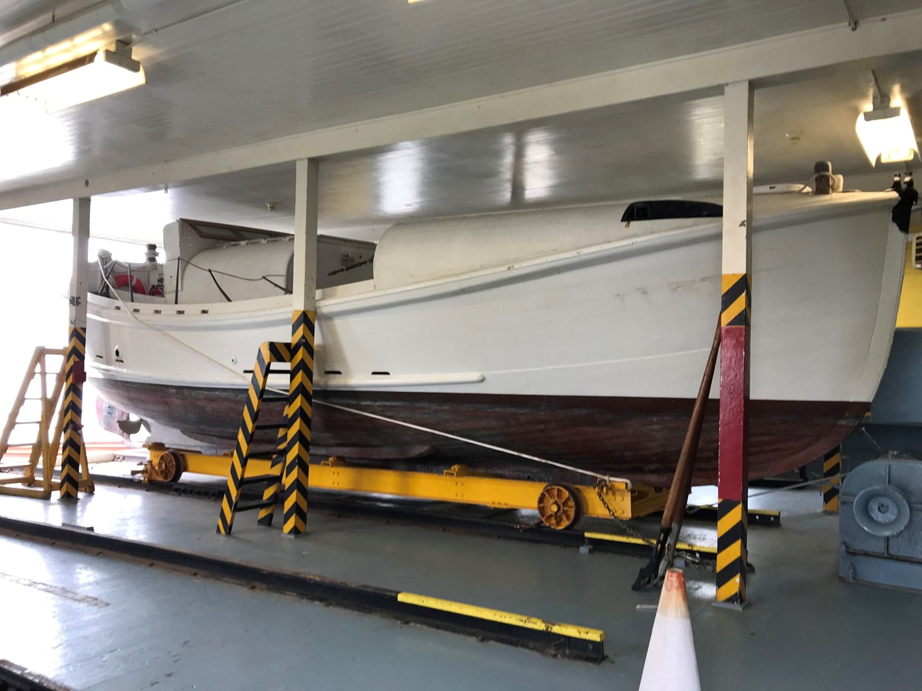 A color photograph of an old, 36-foot-long, motorized lifeboat resting on a rail car on tracks within the boat bay of boathouse. The lifeboat is painted white above the waterline and brick red below.