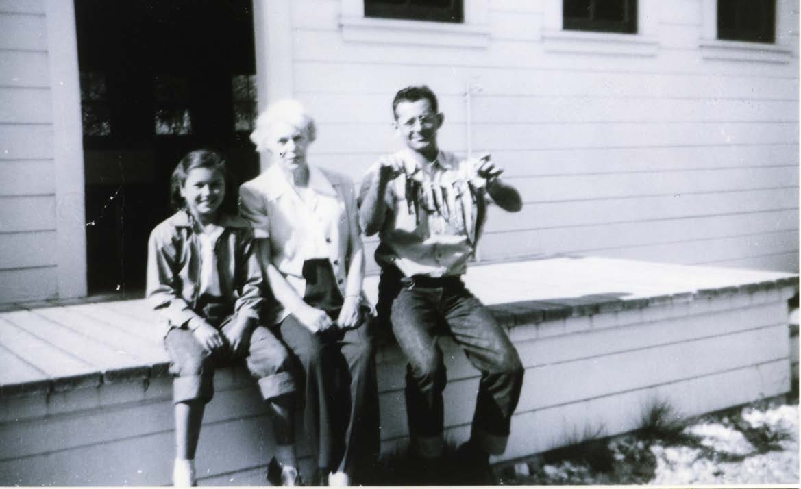 Three people site on a wooden loading dock on a hatchery building. The man is holding fish of varying size on a line.