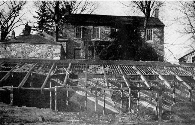 The ruins of a greenhouse stand in front of a two-story stone house. Black and white photo