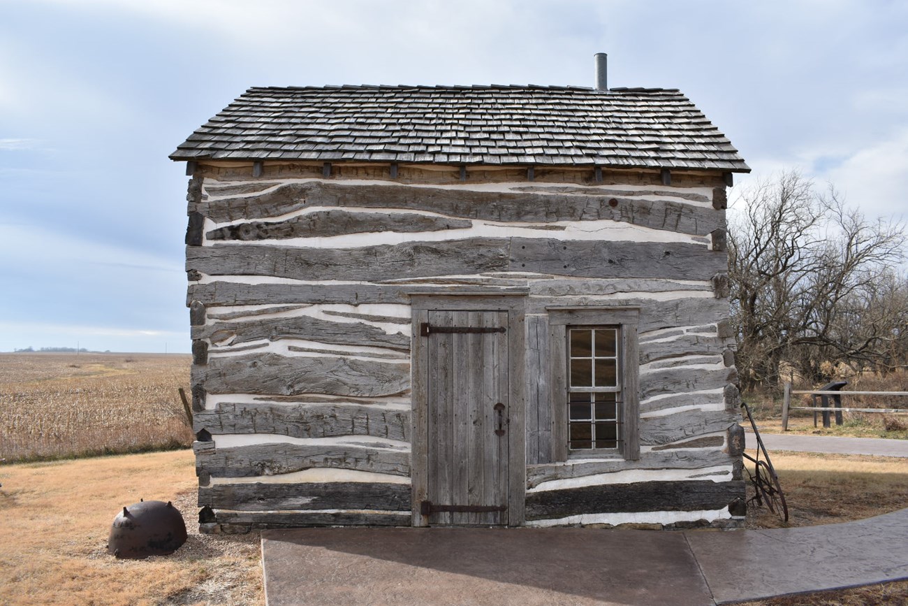 A door and a window in one side of the log Palmer Cabin, with open fields beyond