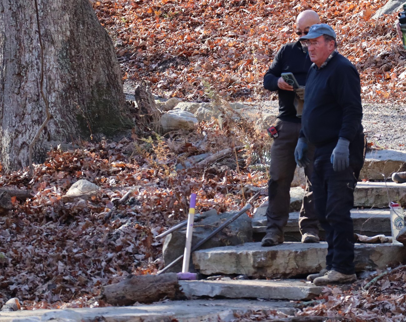 Three people stand on stone steps on a wooded hillside, with several construction tools and work equipment on the ground.