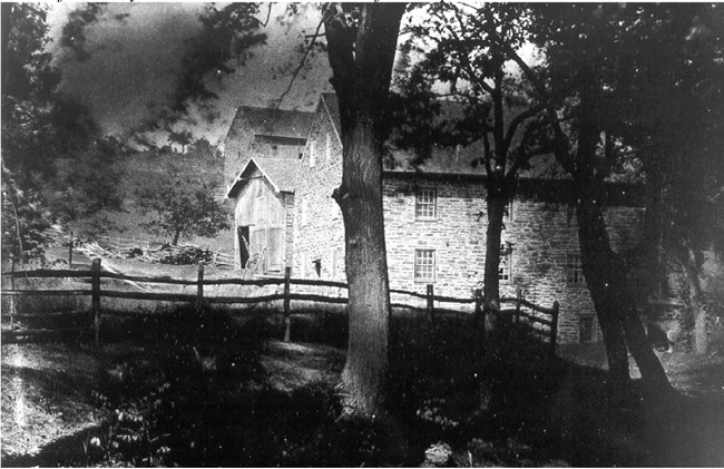 Black and white image of two-story stone mill structure, in an agricultural landscape of fences, outbuildings, and tall trees.