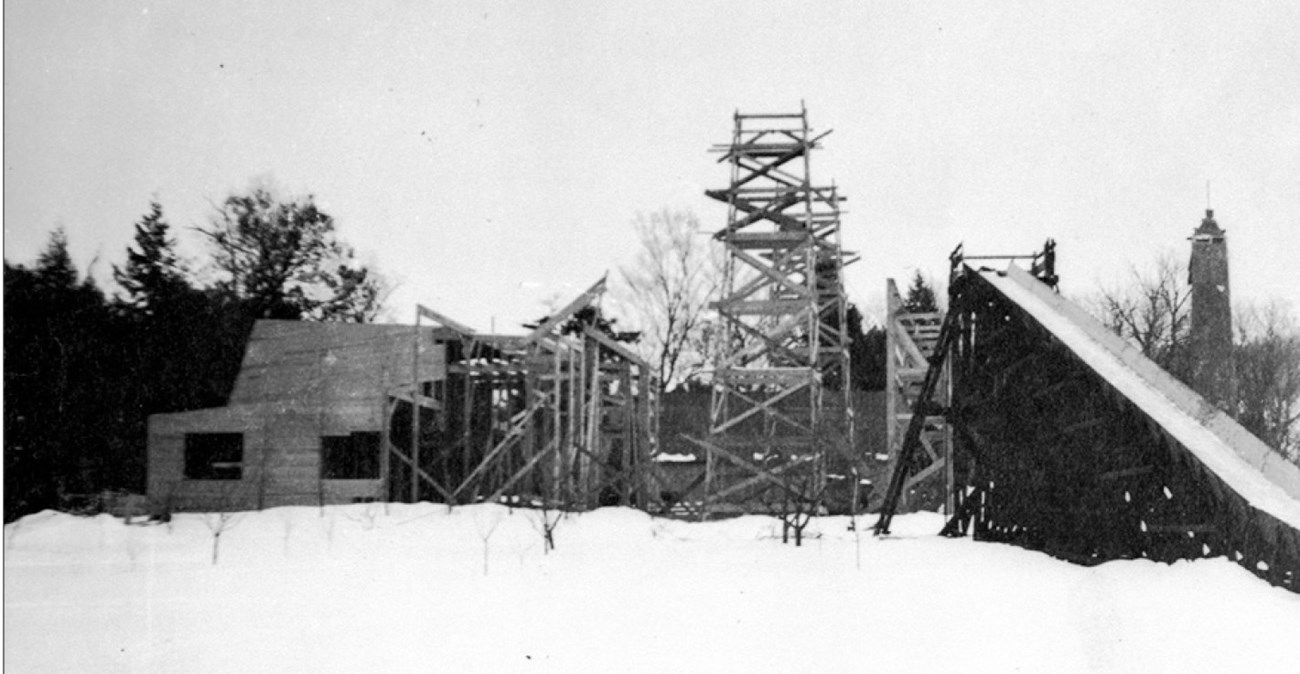 Leafless orchard trees grow in a snowy area in front of a structure under construction. An inclined chute is to the right.