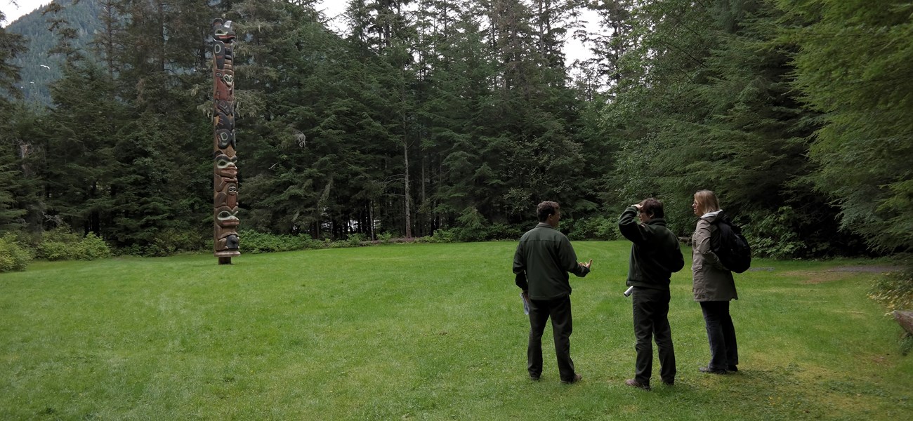 Three people stand in a grassy area facing a totem pole, surrounded by evergreen trees