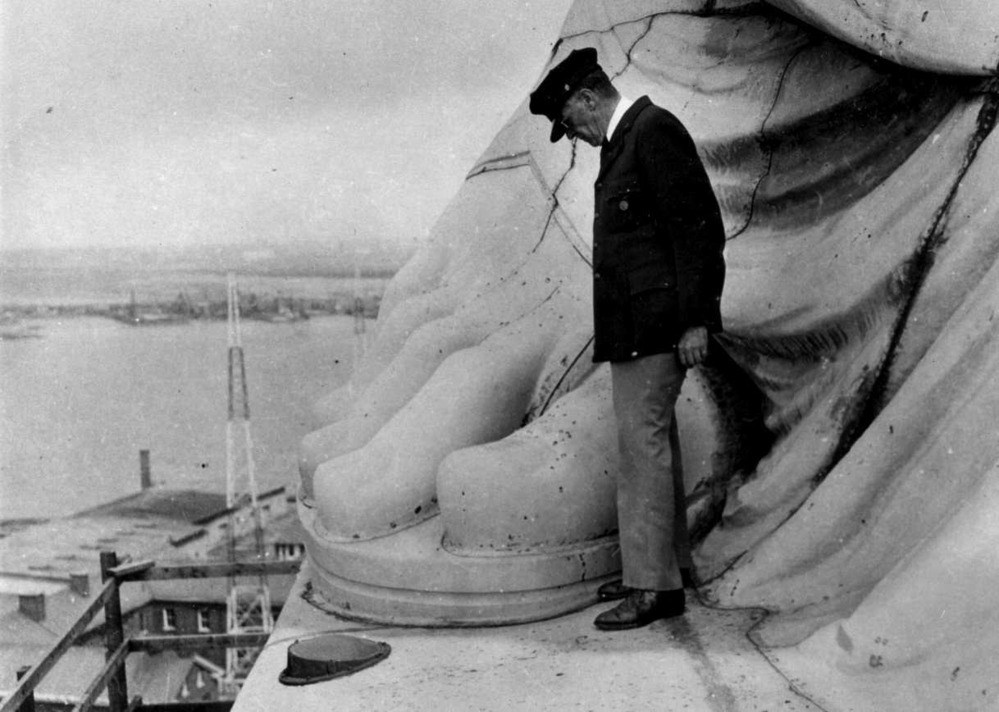 A superintendent with a cap and jacket stands at the base of a a statue beside a large carved foot, overlooking the harbor.