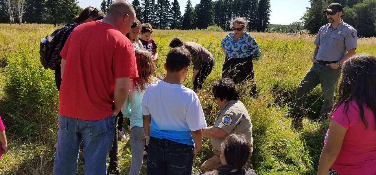 A group of kids and adults crowd over a kneeling guide in a grassy field
