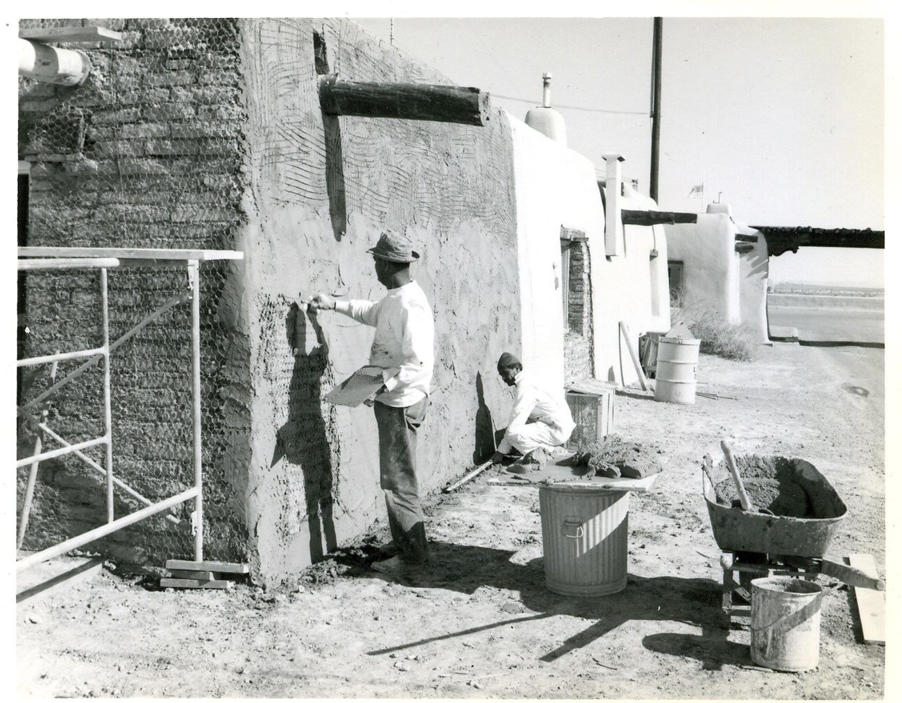 Two people, one standing and one squatting, use tools to finish the surface of an adobe structure.