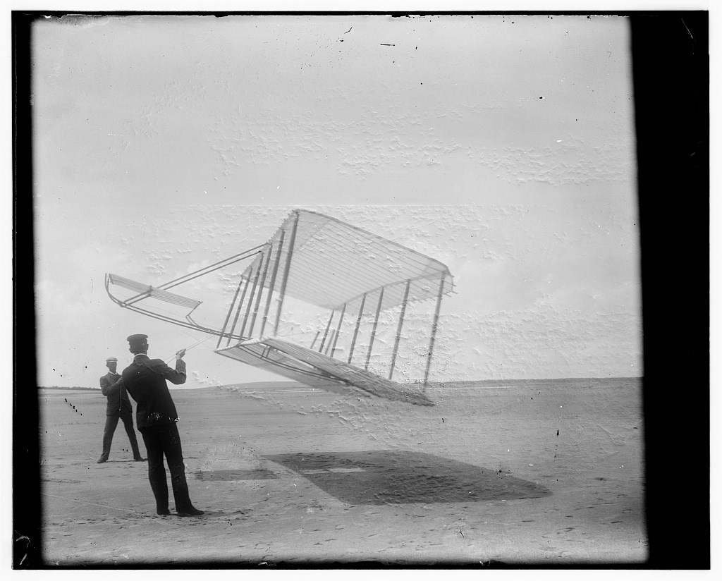 Side view of glider flying as a kite near the ground, Wilbur at left and Orville at right, glider turned forward to right and tipped downward