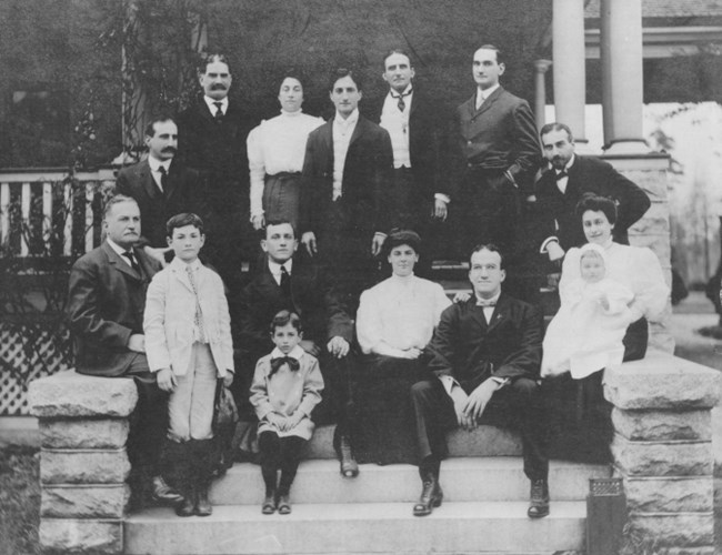 A group of 15 people of varying ages, wearing suits and dresses of 1900, pose on wide stone stairs of a porch.