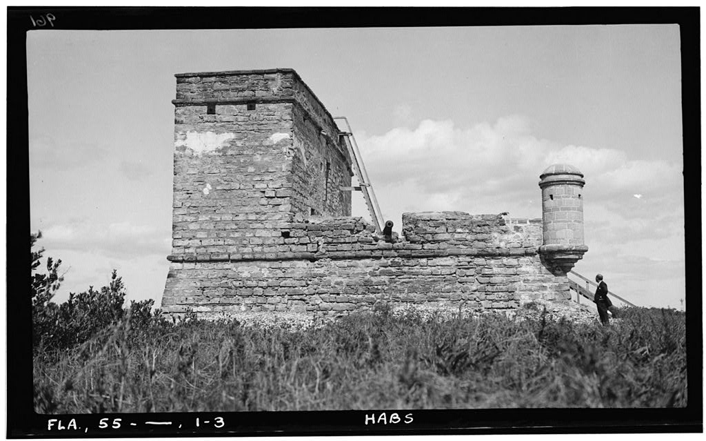 A person stands near the corner of a stone fortification with a rectangular watchtower on one side
