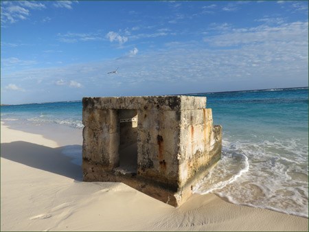 A square concrete bunker with a door on one side is surrounded by a sandy beach and bright ocean water.