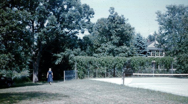 Eleanor Roosevelt walks through short grass beside a tennis court, with tall leafy trees in the background