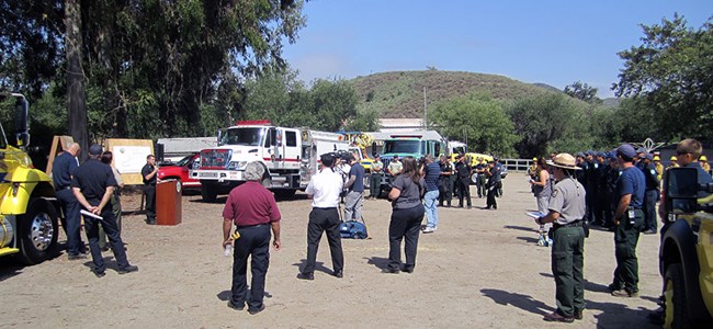 A group of press members and NPS employees stand in an open parking lot during a press conference.