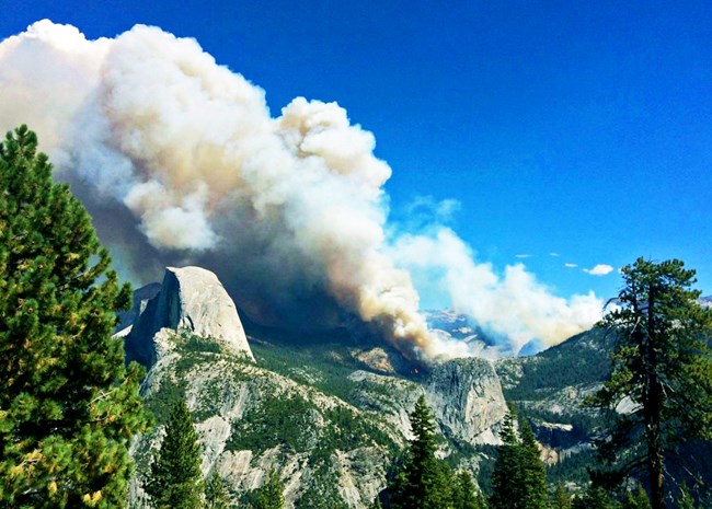 A fire burns in the distance with a left-direction smoke plume. In the foreground is Half Dome.