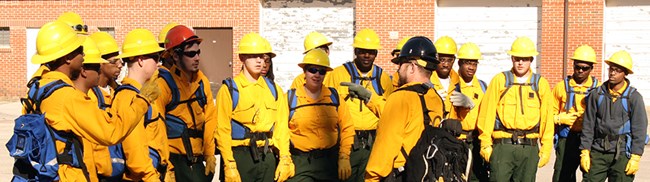 A group of young men and women in firefighting gear listen to a man speaking to them.