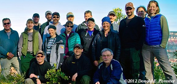 A group is gathered at a viewpoint with the vista of the Grand Canyon behind.