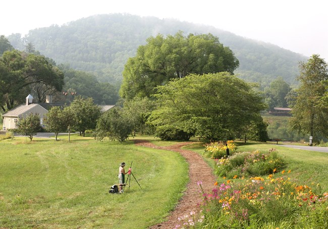 A green field with a dirt road running through it. Mist-covered hills in backgrounds. Green trees. Wildflowers. Someone is painting in center of field.