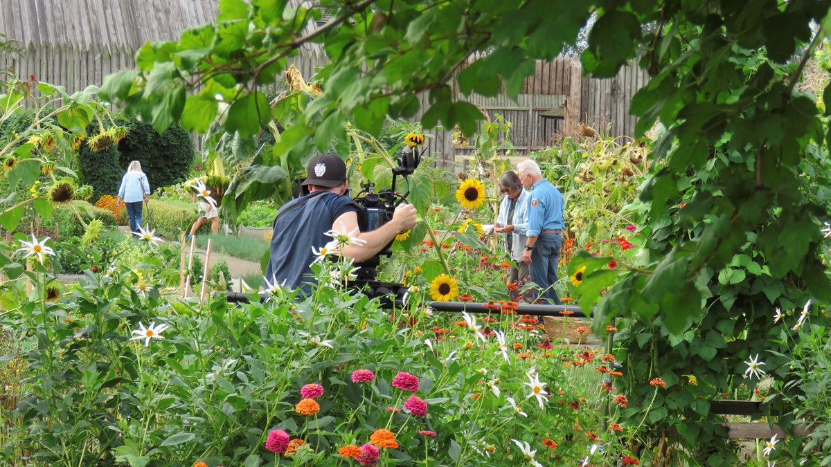 Four people stand in a garden while a man films them with a standing camera