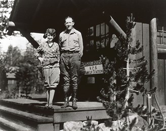 Eva McNally wearing a badge and gun holster stands with husband Charles on the ranger station porch.