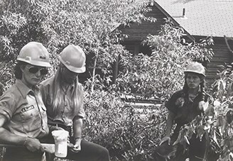 Three women in hardhats stand among plants. The wear the NPS uniform and one holds an axe.