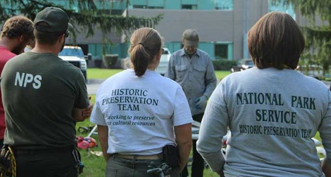 NPS employees standing around an instructor. The back of their t-shirts read "Historic Preservation Team."