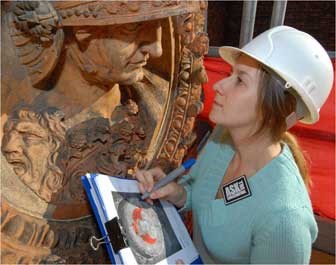 A female intern wearing a hardhat with a clipboard looking closely at a large artifact.