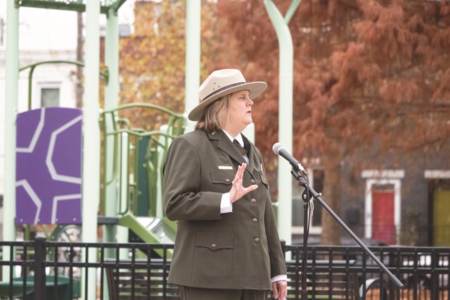 A female ranger wearing a green formal button down uniform and felt flat rimmed hat holds her hand up as she speaks into a microphone with a colorful children’s playground in the background.