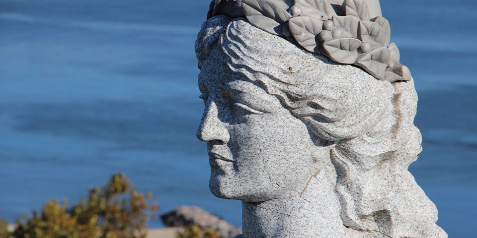 A granite head of a victory, a woman with hair pulled up encircled with a wreath, overlooks blue water.
