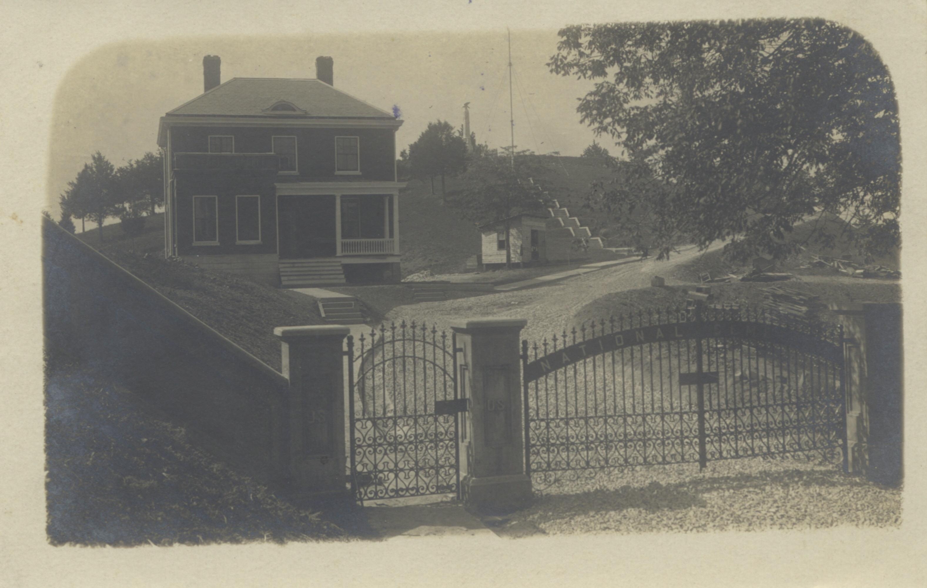 A historic postcard of a two-story lodge to the left of a curving drive, with a metal gate and enclosure wall.