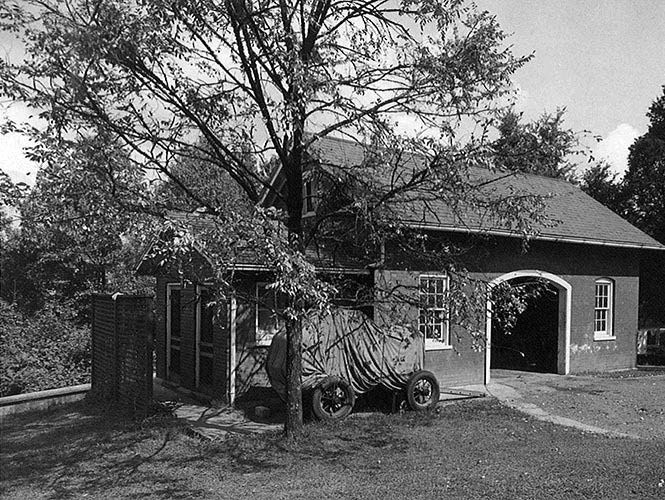 A tarp covered wagon and tree stand beside a brick stable, showing a restroom addition on the side.