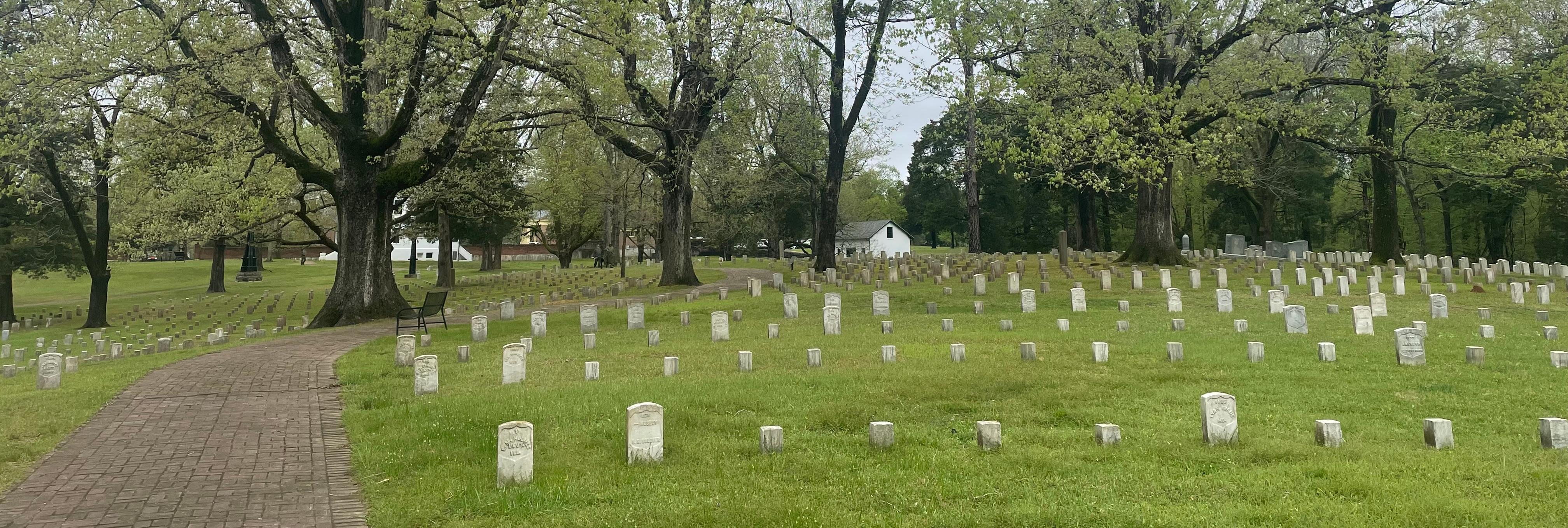 Trees shade a burial area of uniform grave markers alongside a curving brick walkway.