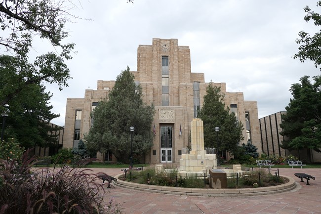 A view of the center of a large light brown building, with a white fountain in front. A clocktower at the center of the building is the highest point in the image.