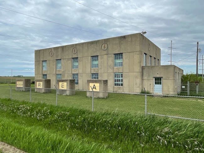 Two-story power plant fronted by four air intake houses in a rural area. There are five large window openings at each level and three circled lightning bolts stamped into cornice.