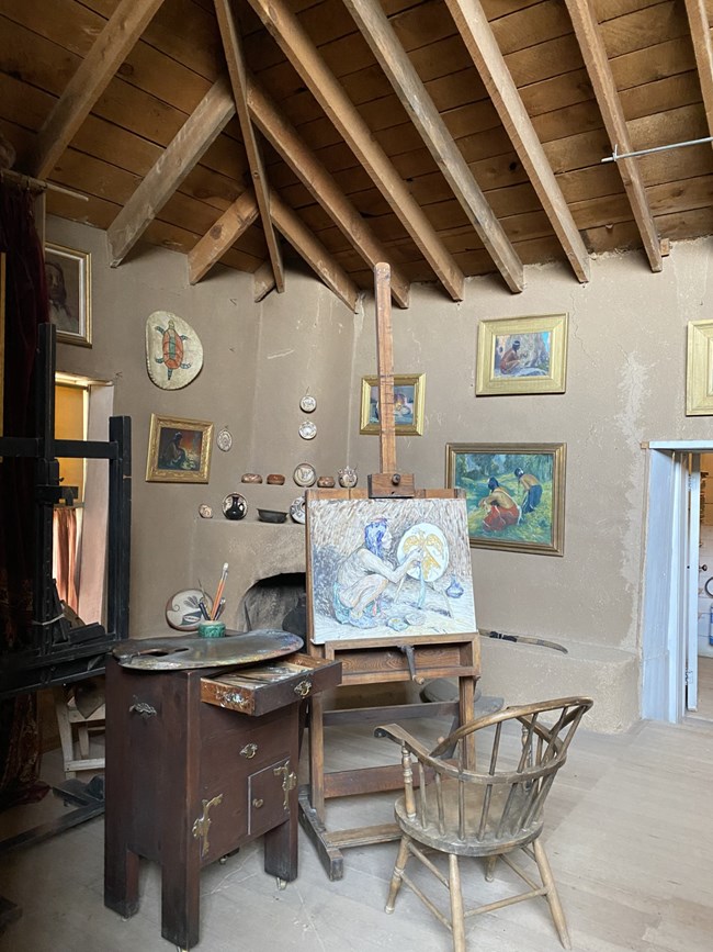 Interior view of a chair seated in front of an easel with a painting. Atleft is a small table with an open drawer and an easel on its surface. In the background is a corner fireplace around which hang framed paintings.