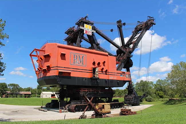 Image of gigantic mining excavator, set among gravel, grass and trees, with smaller equipment in the foreground and buildings in the background.