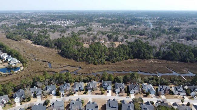 Aerial view of community of houses against a creek, with trees in the background