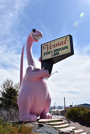 Image of a pink anthropomorphic dinosaur holding a sign, rocks, and plants, with buildings in the background.